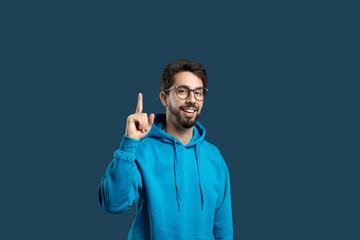 A cheerful young man with glasses and a beard is dressed in a casual blue hoodie, lifting his...