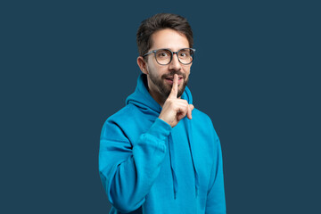 A man in a bright blue hoodie stands against a matching blue background, placing one finger over...