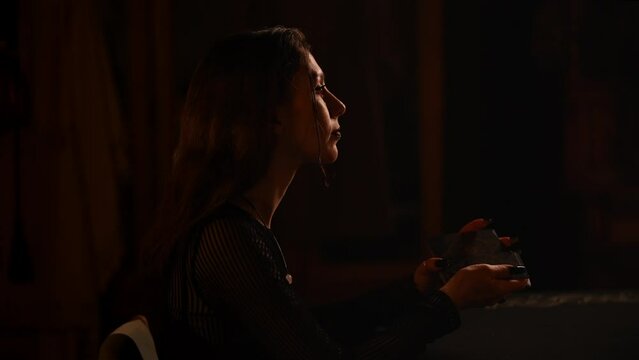 Closeup side view of mysterious fortune teller laying out tarot cards at table in dark room by light of burning candle. Enigmatic fortuneteller shuffling tarot cards, magic ritual, slow motion.