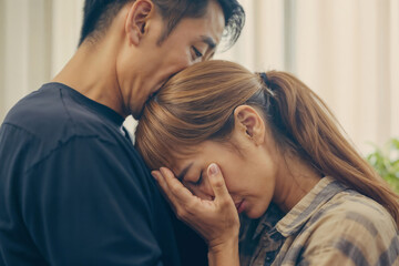 Asian man calms a woman and a woman stands close to each other, a woman cries covering her face with her hand and leans on the man's shoulder on a light blurred living room background - Powered by Adobe