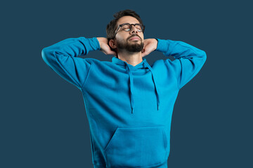 A young man wearing glasses and a casual blue hoodie stands with his hands behind his head, eyes...