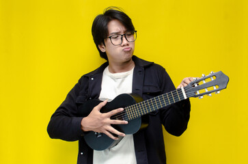 stylish ecstatic asian man playing guitar singing and smiling isolated yellow color background....