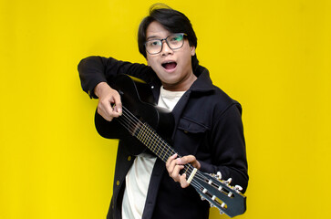 Obraz premium stylish ecstatic asian man playing guitar singing and smiling isolated yellow color background. young asian man plays guitar-ukulele.