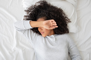 A young woman is lying on a white bedspread, covering her face with her arm as she wakes up. The...