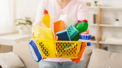 Cropped of woman stands in a sunlit living room, holding a yellow basket filled with various...