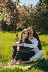 Mother and daughter practicing yoga in a serene, sunlit park, surrounded by lush greenery and blooming trees. The scene captures a warm and peaceful atmosphere, emphasizing health and family bonding