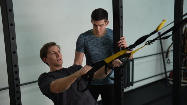 Beginner sportsman doing fitness trx exercises in gym with personal fitness trainer. Male using looped ropes to perform exercises called trx to balance bodies by doing all-body resistance training.