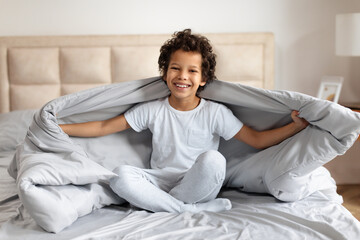 African American young boy with curly hair is sitting on a neatly made bed, his infectious smile radiates joy as he playfully holds up a soft gray blanket in a sunlight-filled bedroom