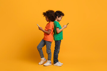 A pair of young African American children stand back-to-back, each absorbed in a smartphone. They...