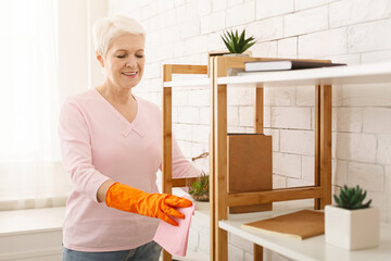 A cheerful elderly woman with short white hair is wearing orange gloves while wiping dust off her...