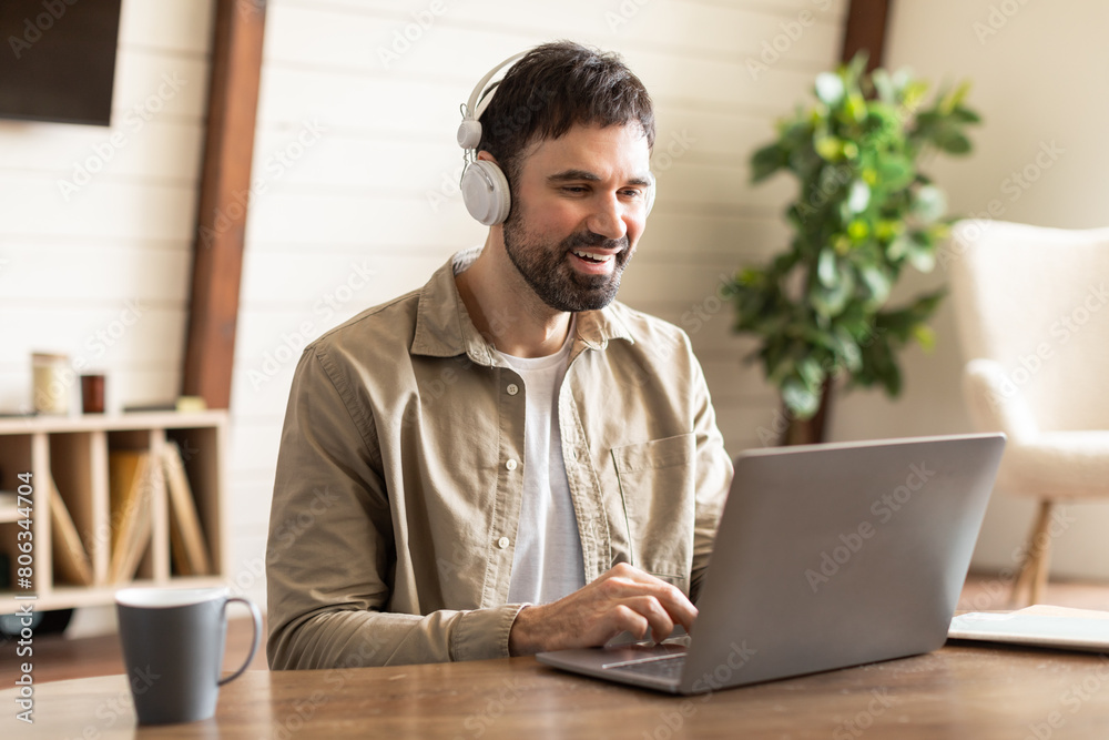 Poster A man is seated at a table with a laptop open in front of him. He is wearing headphones and appears focused on the screen, possibly working or listening to music online