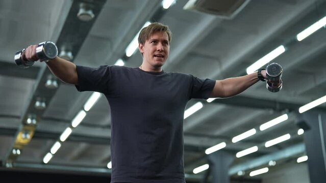 Portrait of motivated beginner sportsman hard time doing lateral arm raise exercise with dumbbells. Handsome male athlete improves shoulders with dumbbell breeding. Shooting in slow motion.