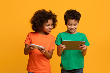 Two young African American children, a boy and a girl, stand side by side, engrossed in the screen of a tablet. They appear focused and curious, exploring the digital world together