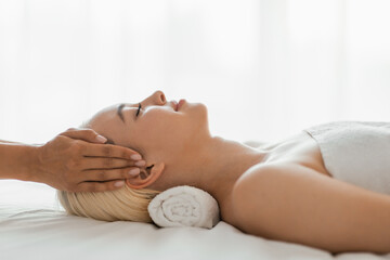 A woman lying down with her eyes closed as a professional masseuse massages her face, focusing on her forehead, cheeks, and jawline, side view