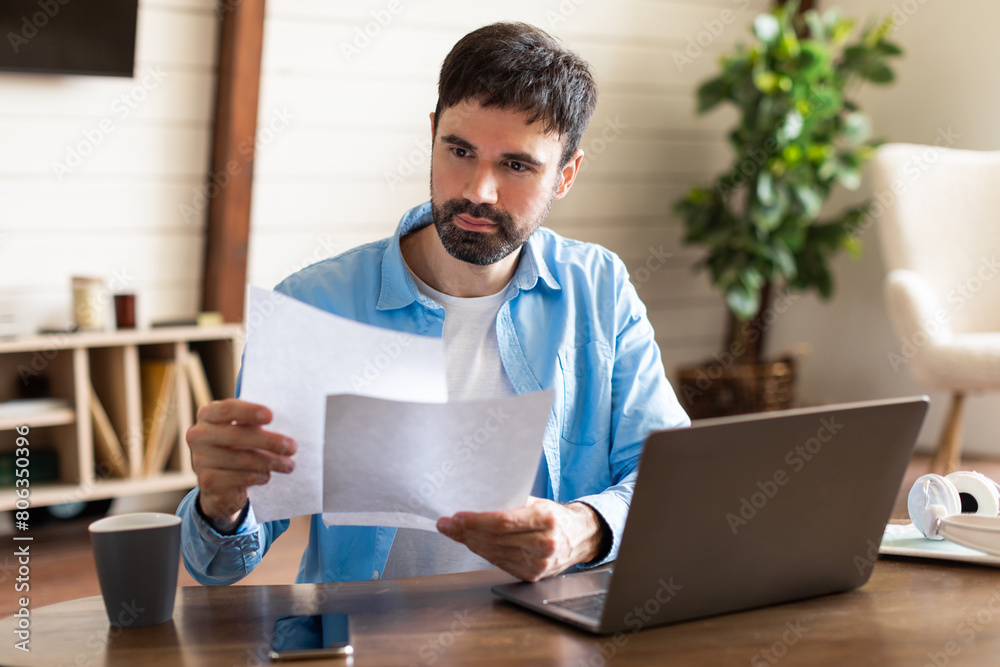 Aufkleber A man is standing and holding a piece of paper in front of a laptop. The man appears focused while looking at the laptop screen and holding the paper