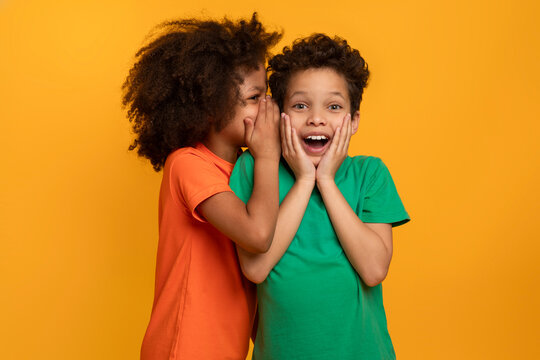 African American cheerful girl whispers into the ear of an excited boy, who reacts with hands on cheeks against a vivid yellow backdrop. Their expressions convey the intrigue of shared secrets.