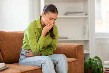 A woman clad in a green shirt and jeans is sitting on a brown sofa, looking distressed with her...