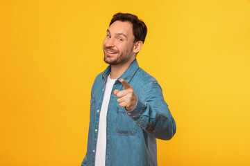 A cheerful man wearing a casual blue denim shirt stands against a vibrant yellow backdrop, pointing...