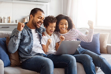 Black family is seated on a couch. They are closely gathered around a laptop, all looking at the...