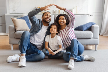 A joyful African American family sits closely on the living room floor with a sofa in the background. The smiling man and woman raise their arms to form roof above their child - Powered by Adobe