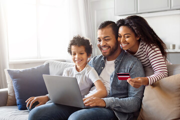 African American family seated on couch, is making an online purchase using laptop and credit card. The parents are guiding son through the process of inputting payment information on the screen.