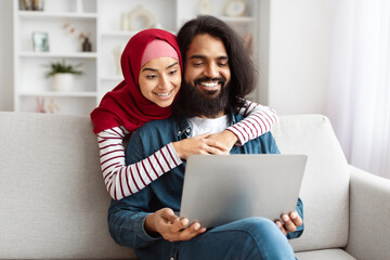 Joyful young Eastern couple engaged in looking at a laptop screen, sharing a moment of laughter in a cozy home environment
