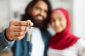 A cheerful eastern man with a beard proudly holds up a set of keys, symbolizing homeownership,...