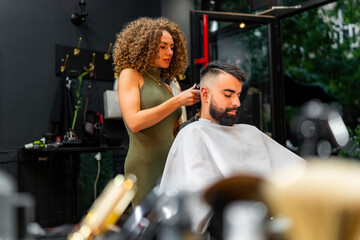 Female barber meticulously trims and styles a male client's hair with electric clippers in a modern barber shop, ensuring a precise and stylish cut. - obrazy, fototapety, plakaty