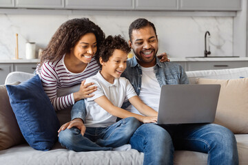 African American family is seated on a couch, all attentively looking at a laptop screen in a...