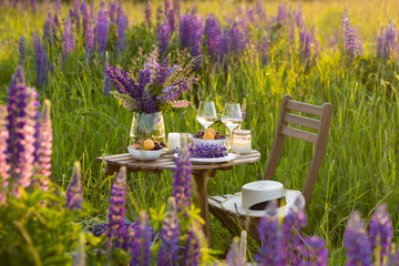 Private wedding party for two, table set with floral decor and greenery, fruits, wine in lupine purple field. Summer, sunset, golden hour. Romantic surprise, marriage proposal. Elegant decoration