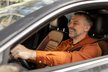 Side view of smiling European man sitting in car on driver's seat, positive driver riding in the city holding hands on steering wheel