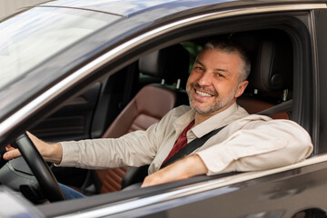 Cheerful middle aged man driving auto, looking through open window and smiling at camera, happy...