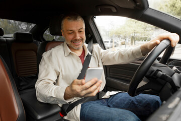 Cheerful middle aged driver man texting on smartphone while driving, sitting in car inside, male...