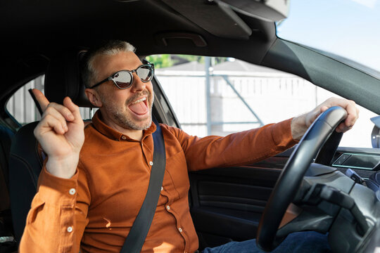 Overjoyed middle aged man in casual outfit enjoying car ride alone, listening to music, singing songs and dancing while driving auto