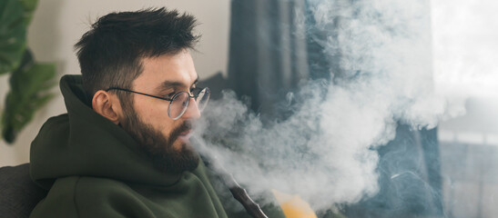 Banner bearded millennial or gen z man smoking hookah while relaxing on sofa at home copy space -...