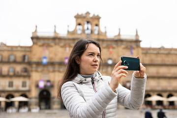 Young adult woman tourist standing with phone making photo or video in old center of spanish city...