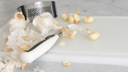 Peeled garlic, garlic press, and kitchen knife close-up on a white plastic cutting board