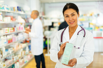 Asian female doctor in white gown standing in drugstore and holding pharmaceutical package in...
