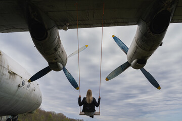 A blonde girl rides on a swing that is attached to the wing of an old An 12 cargo plane. View from the back.