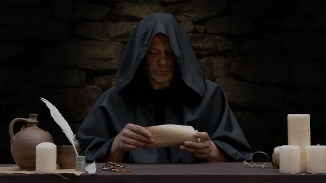 An old monk opens a scroll and reads, sitting at a table against a stone wall, candles burning.