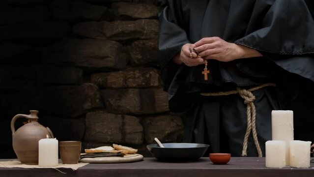A monk in a black cassock sits at a table in a stone cell and eats soup with bread and onions and drinks wine.