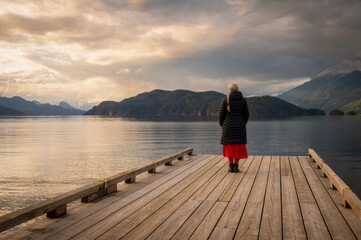 Senior woman gazing out over a lake at sunset from a boat dock. Seen at Harrison Lake, British...