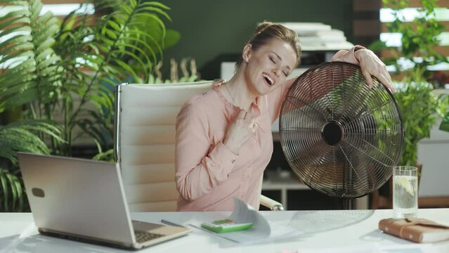 Sustainable workplace. smiling modern middle aged accountant woman at work with electric fan and laptop.