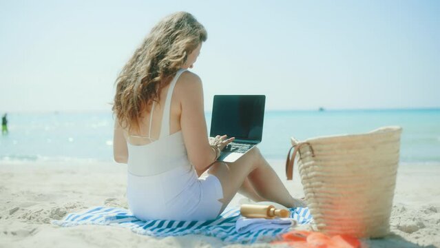 Seen from behind modern woman on the ocean coast with straw bag, laptop blank screen and striped towel.