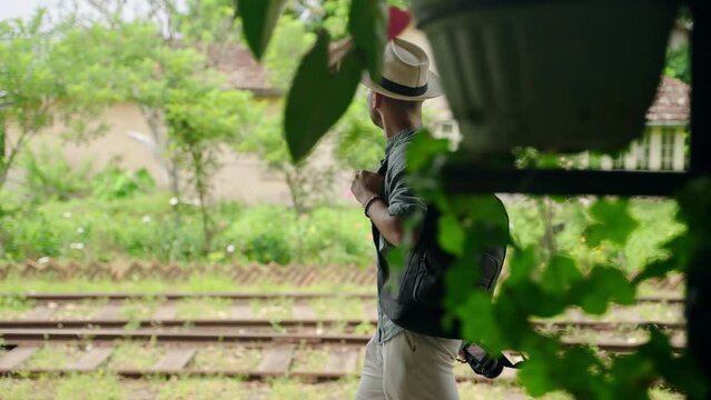 Asian male tourist with camera walks along Haputale railway station. Man explores Sri Lanka culture, nature, wears hat, holds backpack. Solo traveler on adventure, enjoys exotic trip scenery. Slowmo