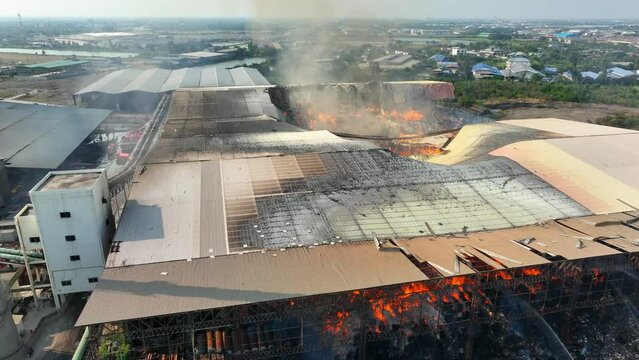 A bird's-eye vista reveals a relentless inferno engulfing an industrial complex. Flames lick hungrily at debris, painting the scene with a fiery hue. Smoke rises ominously, veiling the devastation.
