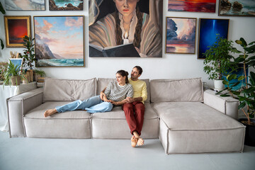 Relaxed creative loving couple sits on couch looking aside window tender hugging at home art...