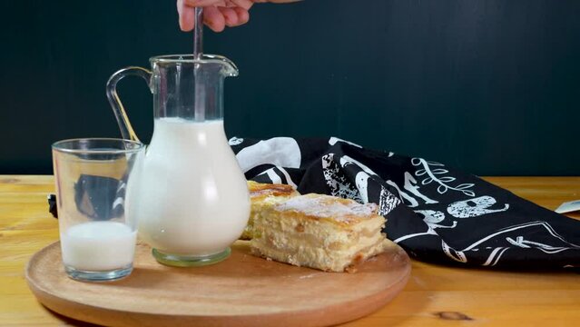 Milk is stirred in a jug, a half empty glass aside, breakfast with freshly baked cottage cheese pie on wooden plate with napkin, food styling on dark background