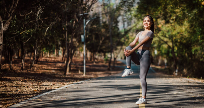 Radiant woman engaged in a standing leg stretch, warming up for her fitness routine on a picturesque forested road.