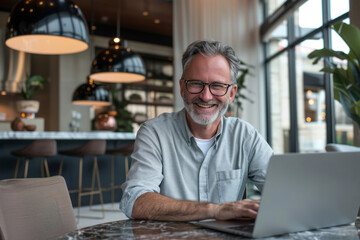 A cheerful middle-aged man with glasses sitting at a table, smiling as he looks into his laptop while typing on the keyboard, his gray hair adding to his charm.